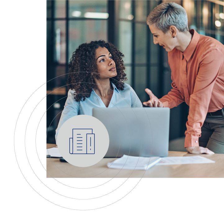 Two women in an office setting at a wooden table with a laptop and papers, engaged in discussion. A document icon overlays the scene, highlighting their task